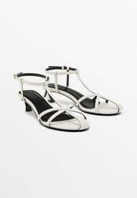 Sandali bianchi in pelle con tacco basso, dotati di cinturini incrociati e una cinghia alla caviglia. L'interno è nero. Texture liscia, design open-toe.