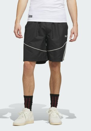 Mann trägt schwarze Shorts mit weißen Paspeln, weiße Turnschuhe, schwarze Socken mit roten Streifen und ein weißes geripptes Tanktop.