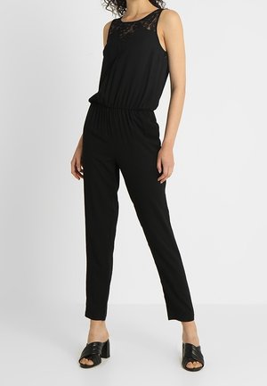 Ærmeløs sort jumpsuit med blonde detaljer ved halsudskæringen, elastisk talje, lige ben, båret med sorte hælede sandaler.