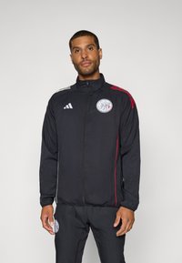Veste de sport noire avec accents blancs et rouges, fermeture éclair, col montant et un logo circulaire sur la poitrine. Tissu léger.