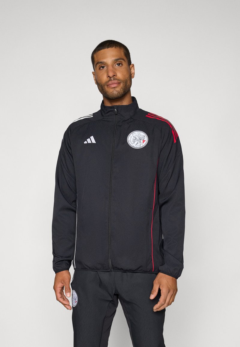 Veste de sport noire avec accents blancs et rouges, fermeture éclair, col montant et un logo circulaire sur la poitrine. Tissu léger.