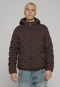 Mørkebrun dunjakke med hette, med en myk, polstret tekstur, frontglidelås og elastiske mansjetter. Brukt over denimjeans.