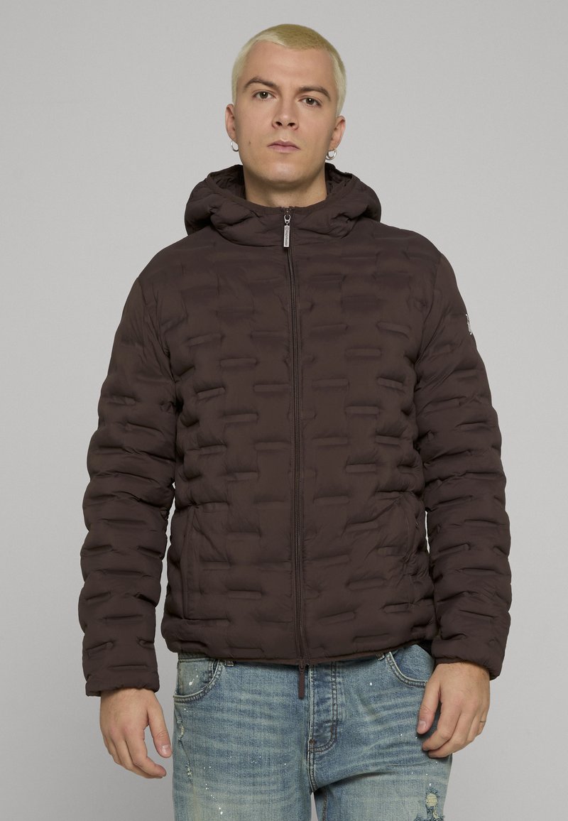 Mørkebrun dunjakke med hette, med en myk, polstret tekstur, frontglidelås og elastiske mansjetter. Brukt over denimjeans.