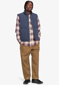 Padded navy vest over a plaid shirt with shades of purple and cream. Loose khaki pants and black shoes with subtle detailing.