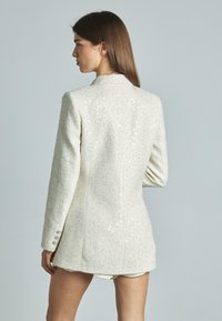 Blazer bianco con paillettes, tessuto texture, chiusura a bottone singolo, maniche lunghe con bottoni decorativi, silhouette leggermente aderente.
