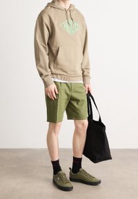 Felpa con cappuccio beige con logo verde, abbinata a pantaloni corti verdi, calze nere, sneakers verdi e una borsa tote nera. Sfondo neutro.