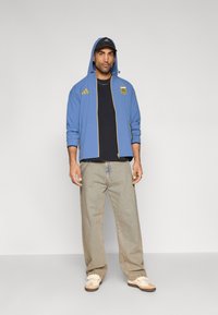 Μπλε αθλητικό μπουφάν με κίτρινα διακοσμητικά, μαύρο μπλουζάκι, φαρδιά μπεζ τζιν και μπεζ αθλητικά παπούτσια· η κουκούλα φοριέται σηκωμένη και περιλαμβάνει λεπτομέρειες με λογότυπο.