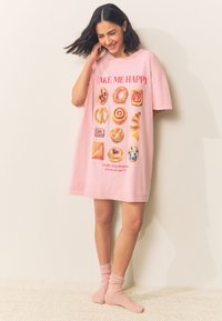 Lichtroze oversized T-shirtjurk met gebakgraphics en tekst: "MAKE ME HAPPY" en "PASTRY OVER PROBLEMS." Zachte stof, korte mouwen.