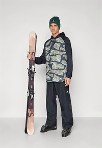 Snowboarder ține schiuri, purtând un hanorac cu model camuflaj, pantaloni bleumarin și cizme negre. Poartă o căciulă verde și legături pentru schi pe schiuri.
