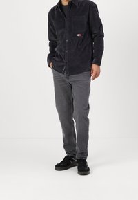 Camicia button-up in corduroy nera con tasca sul petto, abbinata a jeans grigi e sneakers nere con finitura testurizzata.