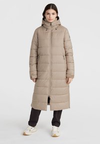Beige lång pufferjacka med huva, vertikalt quiltat mönster, framdragkedja och sidofickor; bars över mörka byxor och sneakers.