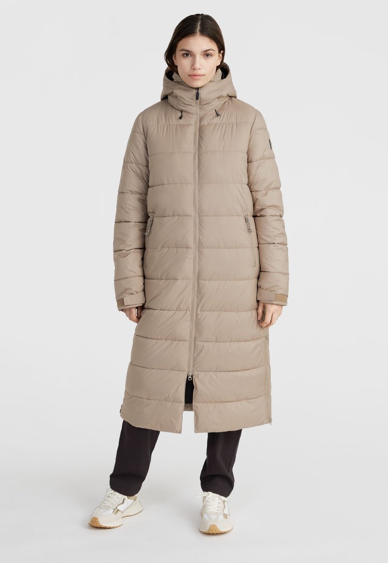 Beige lång pufferjacka med huva, vertikalt quiltat mönster, framdragkedja och sidofickor; bars över mörka byxor och sneakers.