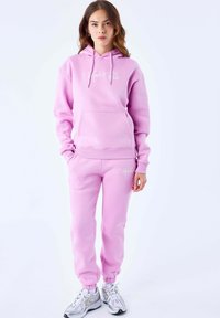 Hoodie rosa con tasca frontale e cordini, abbinato a pantaloni della tuta rosa coordinati con caviglie a costine; accenti del logo bianco visibili.