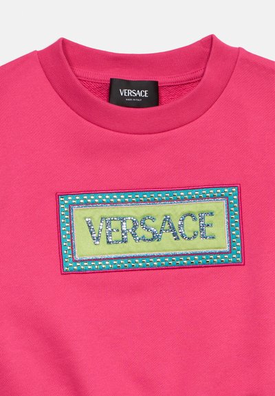 Φούξια φούτερ με πράσινο και τιρκουάζ μπαλώματα που φέρουν το «VERSACE» σε μπλε στρας και με μαύρη ετικέτα Versace στη μέσα πλευρά του γιακά.