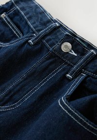 Jean en denim bleu foncé avec un design classique à cinq poches, un bouton en métal argenté et des coutures blanches sur les coutures et les poches.