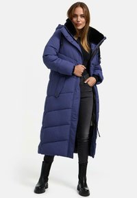 Lungo cappotto invernale blu imbottito con colletto in pelliccia sintetica, caratterizzato da chiusura con zip e bottoni, tasche laterali e cappuccio. Abbinato a jeans neri e stivali.