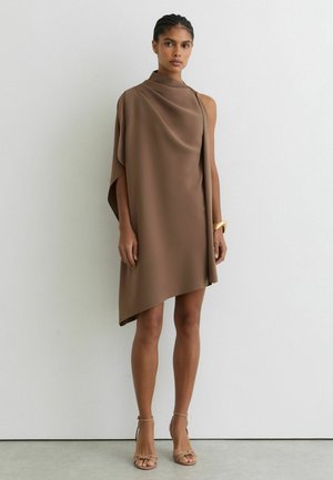 Femme portant une robe taupe asymétrique sans manches avec un tissu drapé, une épaule découverte, un bracelet doré et des sandales à talons hauts couleur chair, debout contre un mur blanc.