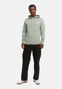 Sudadera verde claro con cordones y un bolsillo frontal, combinada con pantalones cargo negros que tienen bolsillos laterales y zapatillas blancas.