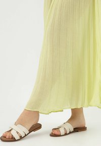 Platte sandalen met een lichtbeige leren bovenkant met gekruiste banden, tegen een doorschijnende, lichtgele stroomlijnrok.