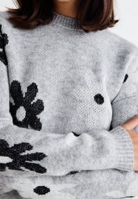 Femme portant un pull en maille gris avec de grands motifs floraux noirs et blancs, bras croisés, montrant une partie de son visage et de ses cheveux.