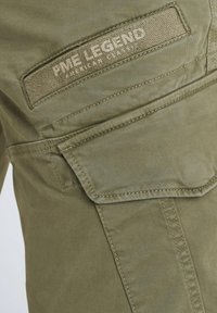 Poche de pantalon cargo kaki avec rabat cousu et étiquette en tissu PME Legend American Classic au-dessus de la poche.