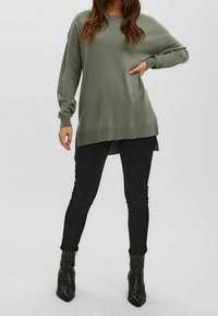 Langärmliger grüner Pullover mit entspannter Passform, gerippten Bündchen und hohem-abfallendem Saum, kombiniert mit schwarzen Skinny Jeans und Stiefeletten.