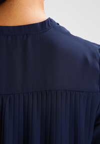 Blusa blu navale, tessuto leggero, presenta pieghe verticali sul retro, con un colletto rialzato e cuciture sottili lungo il colletto.