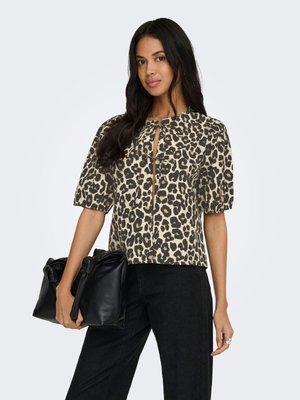 Young woman with long dark hair wearing a beige and black leopard print blouse, black pants, holding a black leather handbag.
