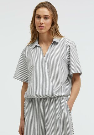 Woman standing with one hand in her pocket, wearing a short-sleeve, collared shirt and pants with thin vertical black and white stripes.