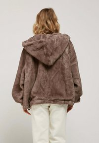 Veste en fausse fourrure marron avec une capuche, coupe oversize, présentant un tissu texturé et un ourlet qui tombe en dessous de la taille. Un pantalon blanc complète la tenue.
