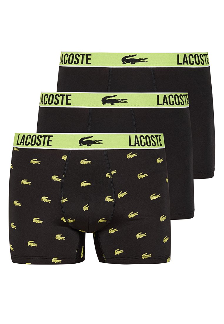 Trzy czarne bokserki Lacoste, dwie gładkie i jedna wzorzysta z żółtymi logo krokodyla, wszystkie z żółtymi gumkami w pasie z tekstem i logo Lacoste.