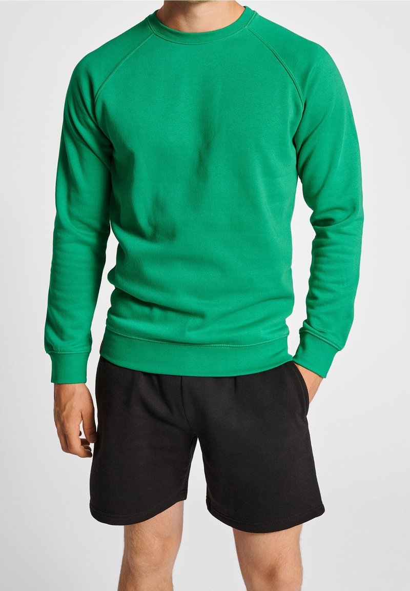 Homme portant un sweat-shirt vert vif à manches longues et un short noir, debout avec une main dans la poche sur un fond uni.