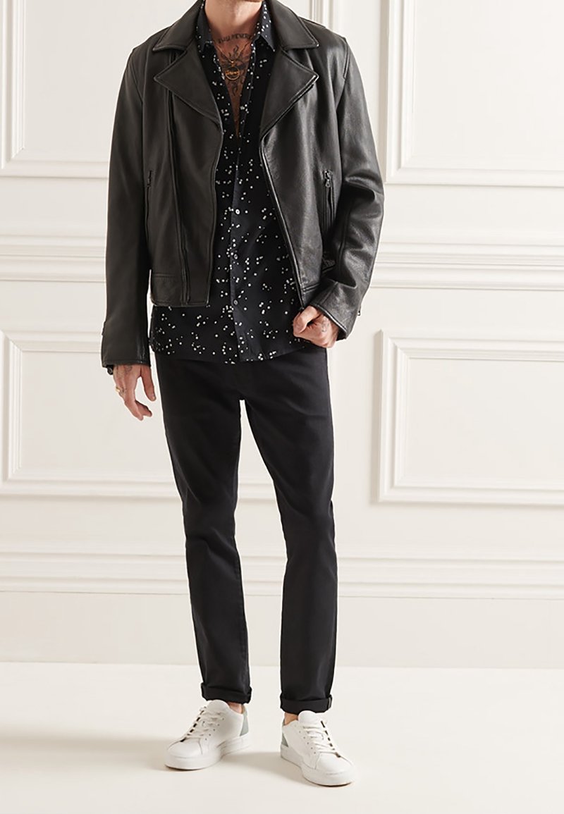 Homme portant une veste en cuir noire, une chemise noire à motifs, un pantalon noir et des baskets blanches, debout contre un mur blanc à panneaux.