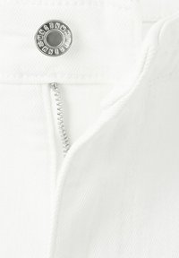 Bela teksas tkanina s delno odprtim srebrnim zadrgo in kovinskim gumbom, vgraviranim z imenom blagovne znamke.