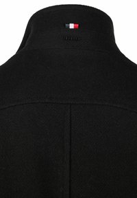 Abrigo de lana negra con un cuello alto, con un logotipo en la parte posterior y una etiqueta tricolor en rojo, blanco y azul. Tejido suave y texturizado.
