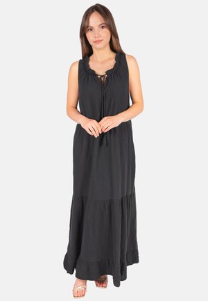 Femme portant une robe maxi noire sans manches avec un col à volants et un détail noué, se tenant debout les mains jointes devant.
