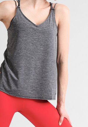 Personne portant un haut sans manches gris et des leggings rouges, debout avec une main posée sur sa jambe pliée.