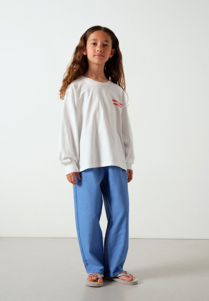 Fille aux longs cheveux bruns portant une chemise blanche à manches longues, un pantalon bleu et des sandales blanches, debout sur un fond clair uni.