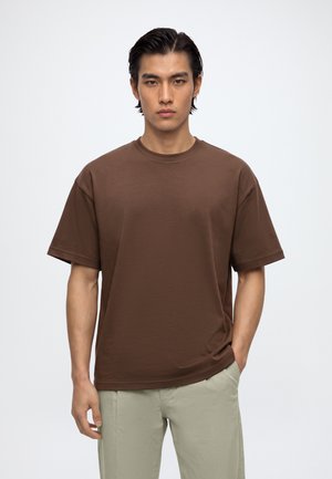 Un homme porte un T-shirt marron à manches courtes et un pantalon clair sur un fond de studio blanc uni.