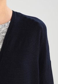 Tummansininen neulecardigan, jossa on V-pääntie, ja joka sisältää tekstuurilla varustetun raidoituksen sekä leveät hihat. Pehmeä, kevyt kangas ja rento istuvuus.