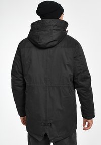 Chaqueta larga negra con capucha, hecha de tela texturizada. Presenta un collar acolchado, dobladillo ajustable y cierre de botones oculto.