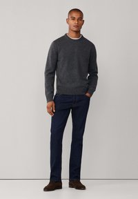 Grijze gebreide trui met geribde manchetten en zoom, gecombineerd met donkere blauwe jeans en bruine schoenen, staand tegen een neutrale achtergrond.