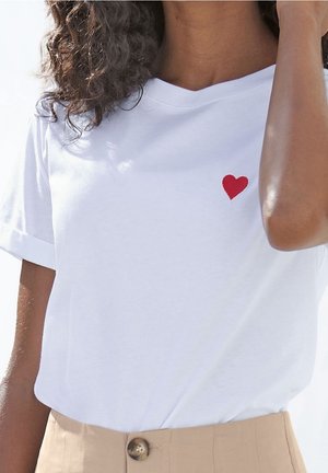 Biała bawełniana koszulka z czerwoną haftowaną serduszkiem na lewej piersi, z krótkimi, podwiniętymi rękawami i luźnym krojem.