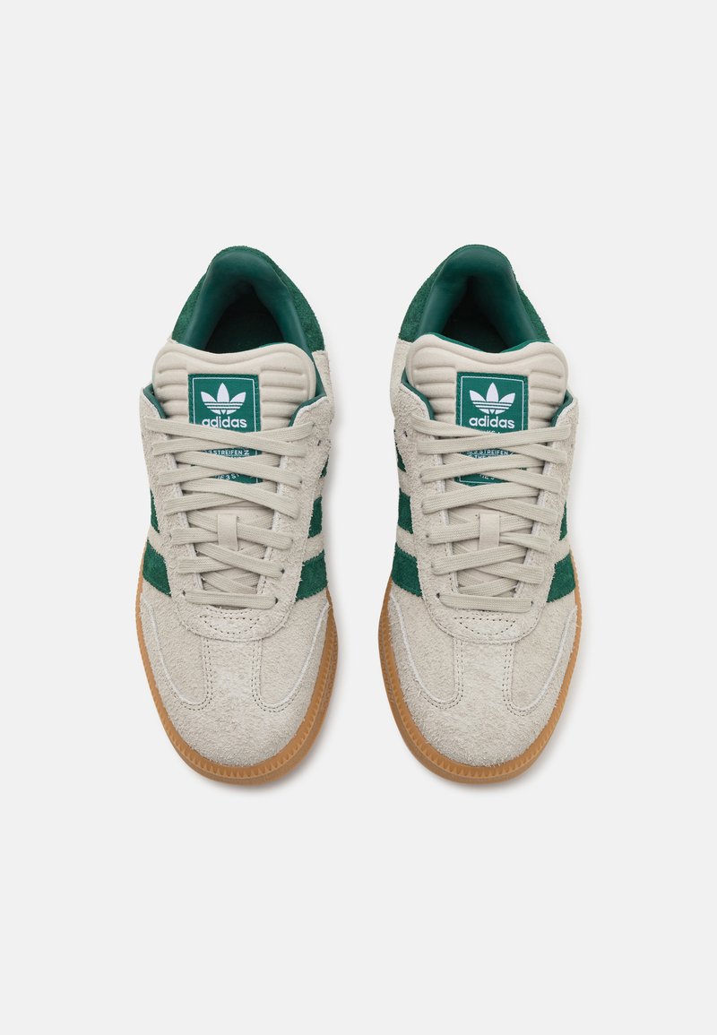 Zapatillas Adidas con parte superior de ante gris, acentos verdes y una suela de goma texturada. El diseño incluye las clásicas tres franjas.