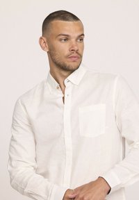 Chemise blanche à manches longues en tissu léger, avec une poche sur la poitrine gauche et une finition légèrement texturée.