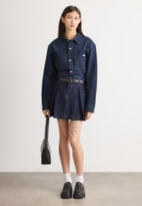 Τζιν πουκάμισο με κλείσιμο από κουμπιά, δύο τσέπες στο στήθος και γιακά, συνδυασμένο με ψηλόμεση πλισέ τζιν φούστα και μαύρα παντόφλια.