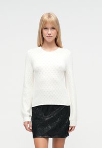 Pull blanc tricoté avec un motif en diamants texturé et manches longues, assorti à une mini-jupe en dentelle noire ornée d'accentuations florales.