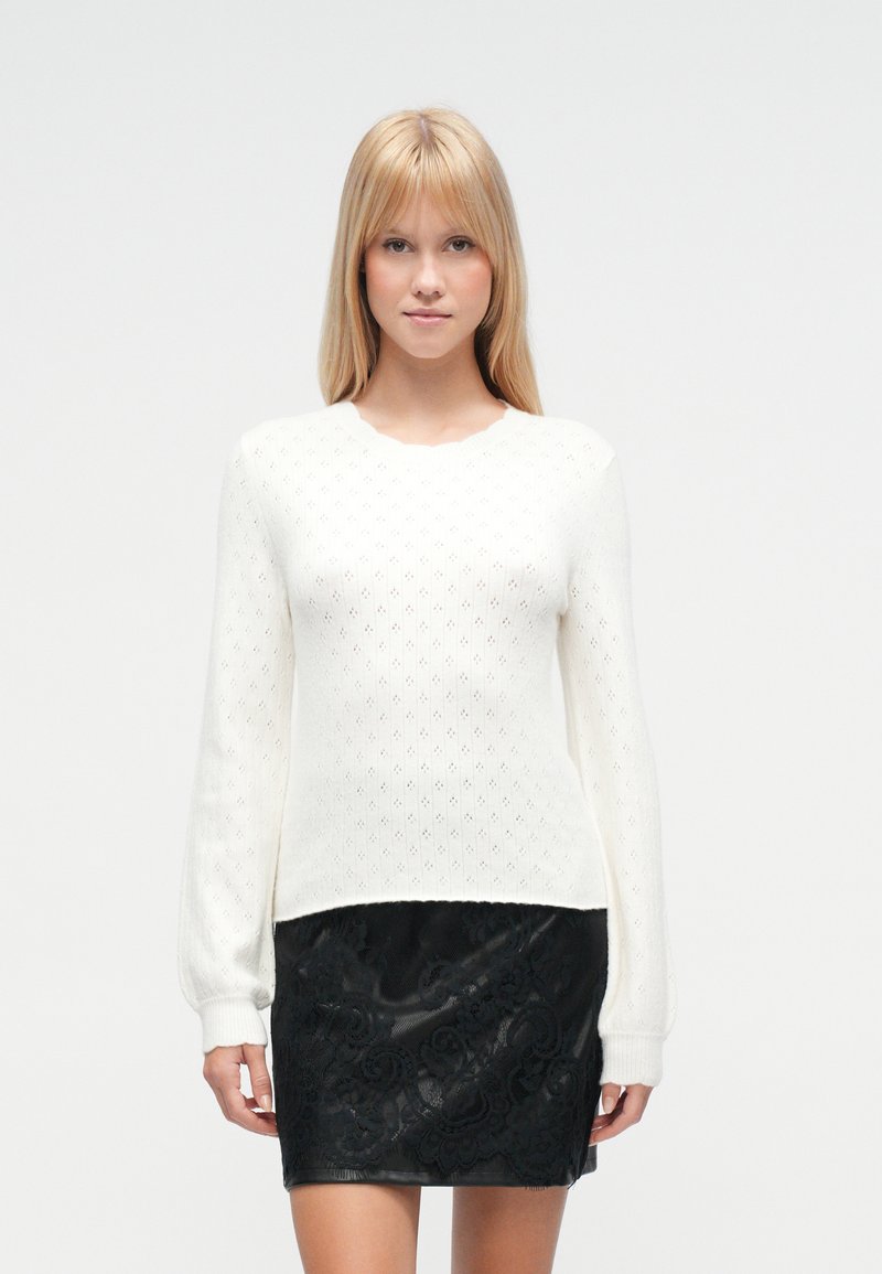 Pull blanc tricoté avec un motif en diamants texturé et manches longues, assorti à une mini-jupe en dentelle noire ornée d'accentuations florales.