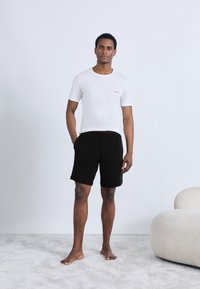 Camiseta blanca de algodón con el logo rojo "HUGO", combinada con pantalones cortos negros de cintura elástica. La modelo está descalza sobre una alfombra gris claro.