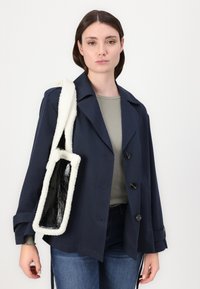 Marineblauwe buitenjas met grote revers en twee voorknopen, gecombineerd met een zwart-witte schoudertas met textuur.